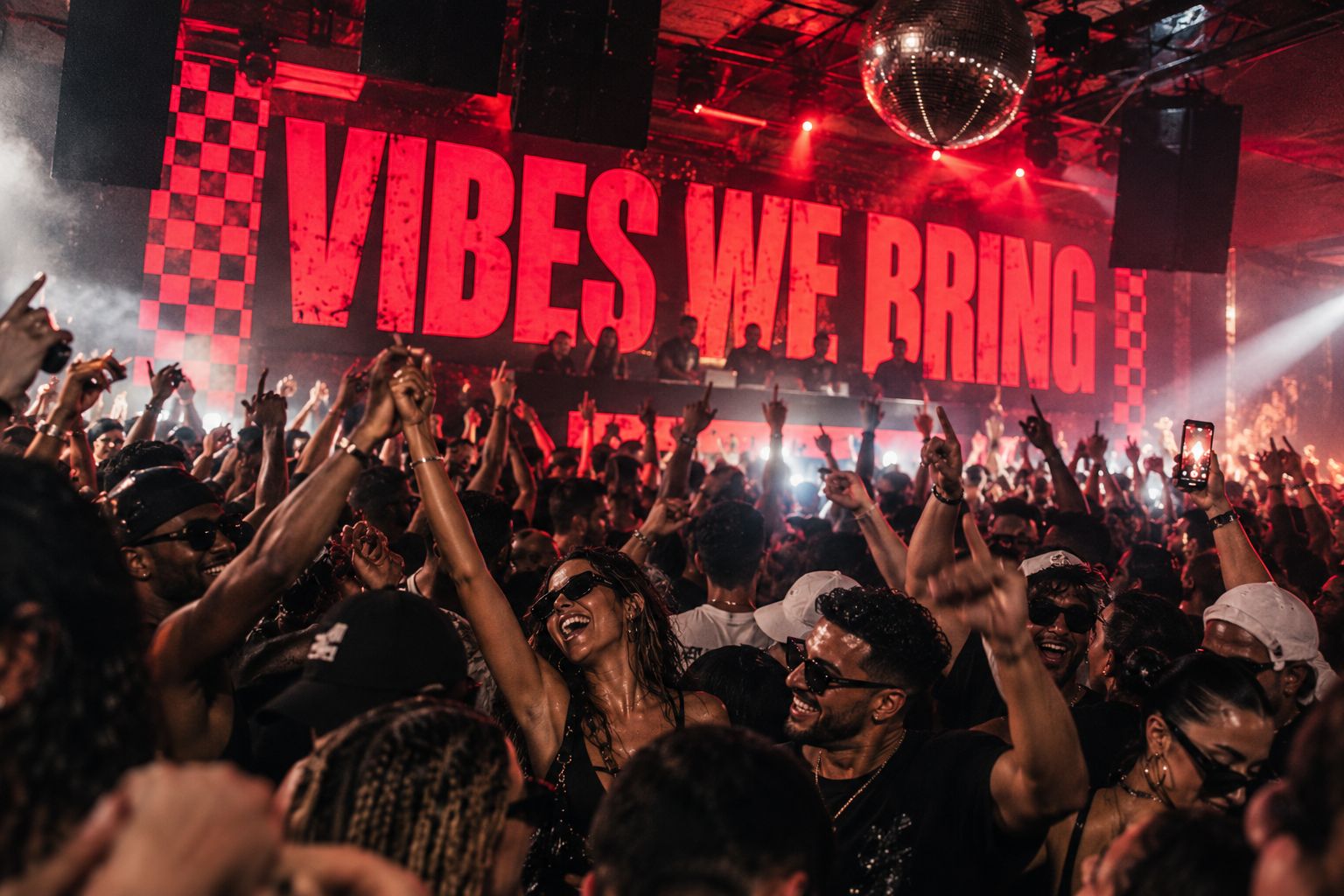 A packed dancefloor with hands in the air beneath a huge red Vibes We Bring screen with checkerboard edges, a DJ booth and a mirror ball above.